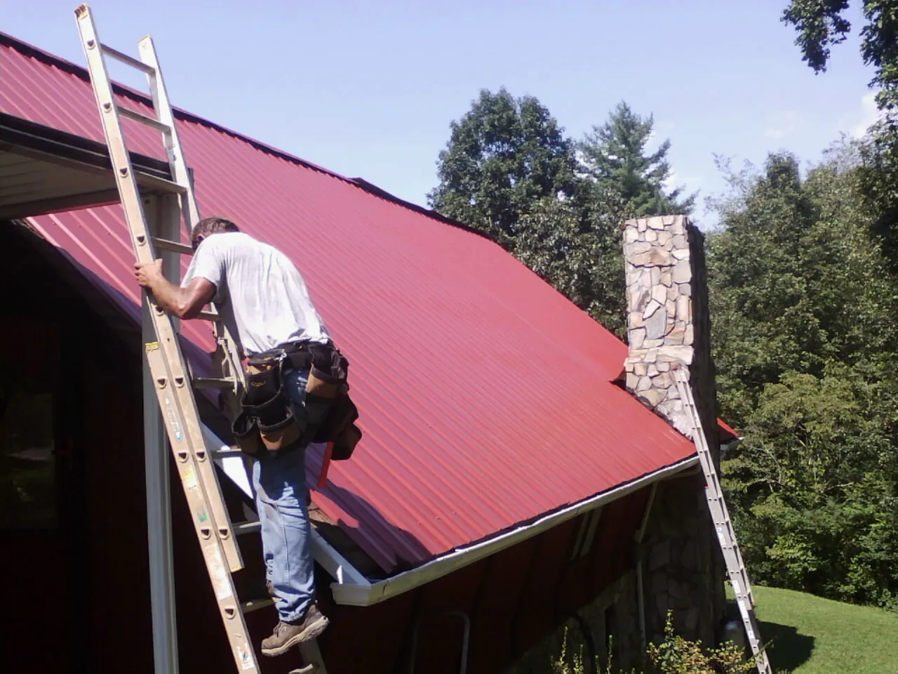 Roofer on a ladder beside a red metal roof and stone chimney — Soffit & Fascia Repair by Roofing Brothers in Swiftwater